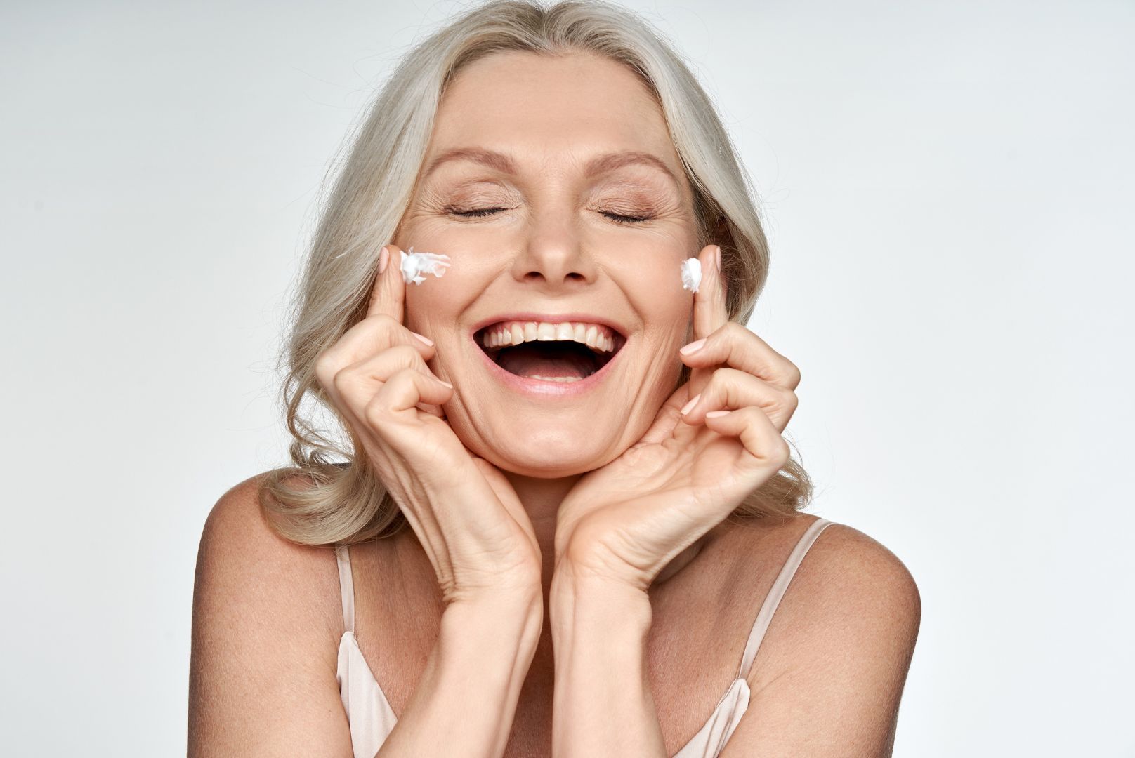 An older woman wearing white camisolesmiling with eyes closed putting cream on her cheeks